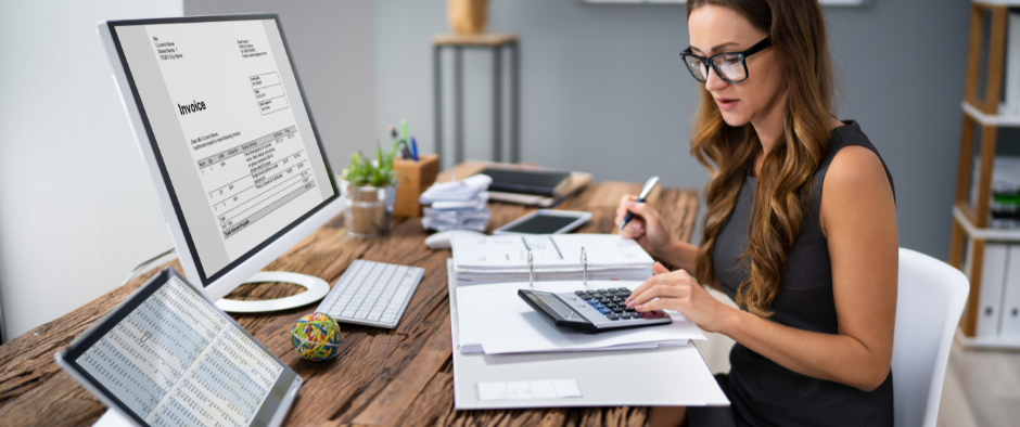 Business owner overwhelmed with paperwork, calculator, and laptop on a desk, symbolizing the hidden costs and stress of DIY accounting compared to the benefits of professional services.